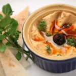 A close-up of creamy homemade hummus with pita slices, garnished with olive and herbs.